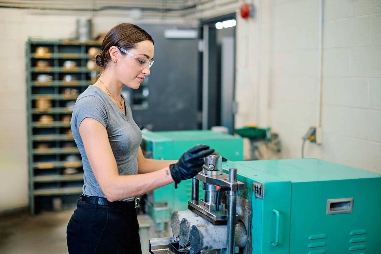 A young woman operating a machine in a workshop, wearing protective gloves and glasses