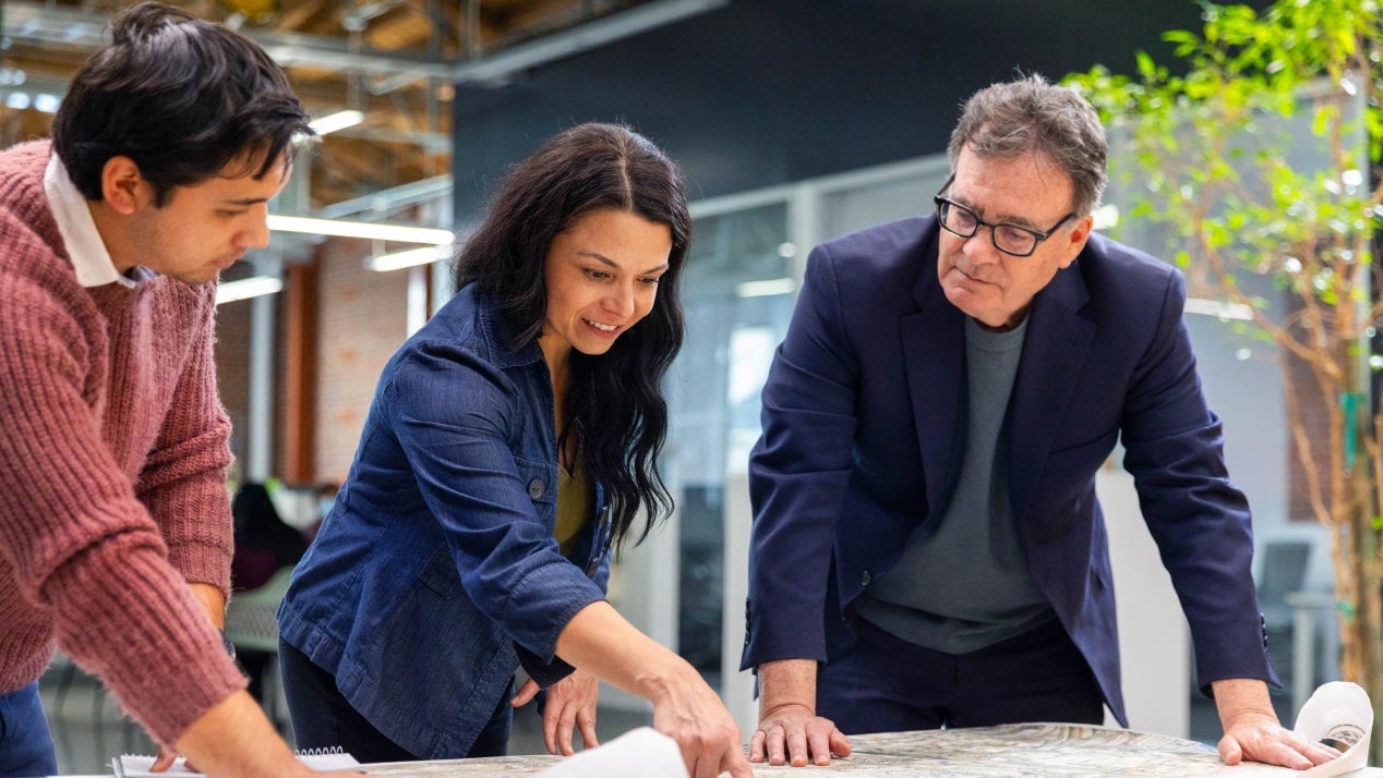 Three workers in business casual clothing discussing a large printed document on a table, presumably a blueprint.