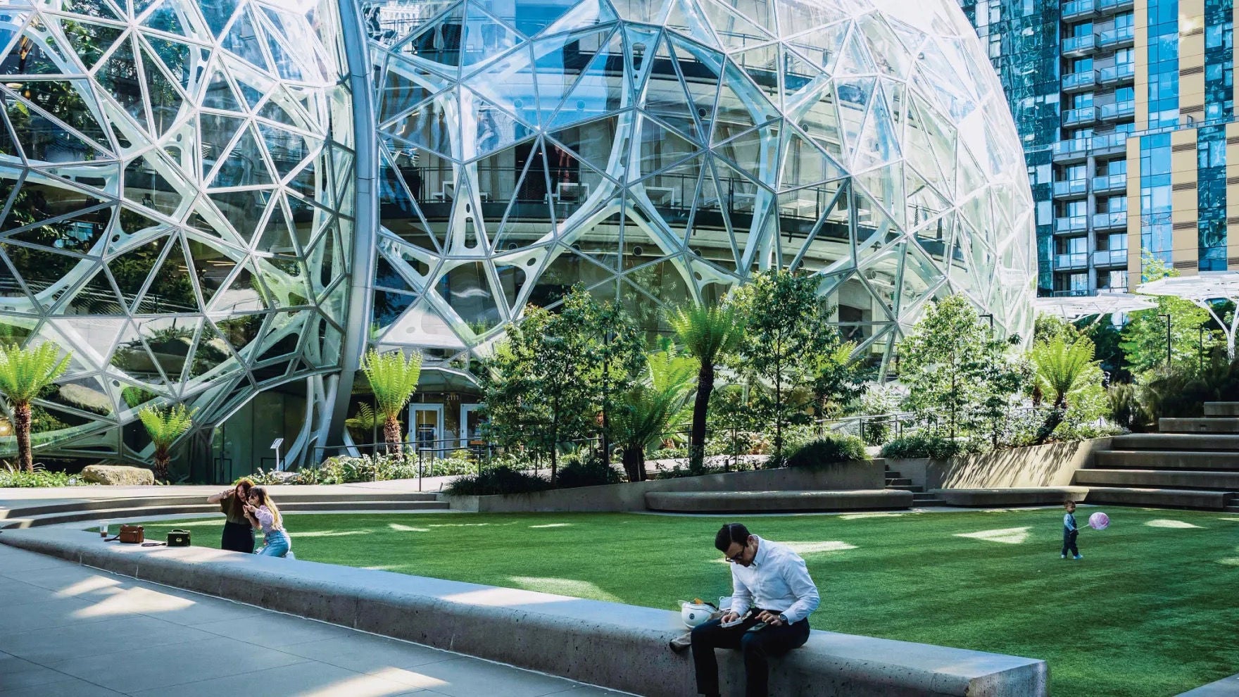 Exterior view of the Spheres from an adjacent plaza, where various people are at leisure amongst a grass lawn and rich landscaping. 