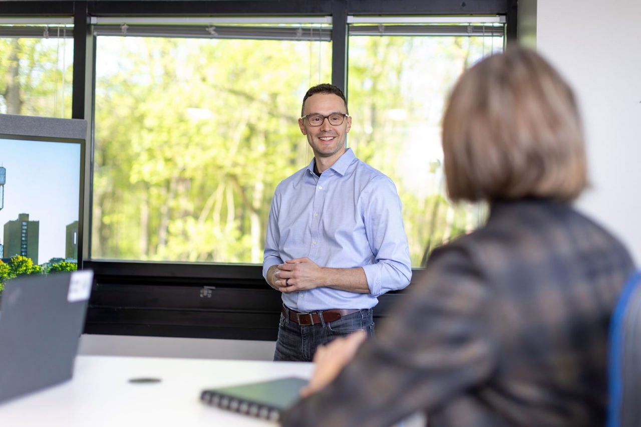 Man in blue shirt smiling and speaking to a seated woman in a bright office.