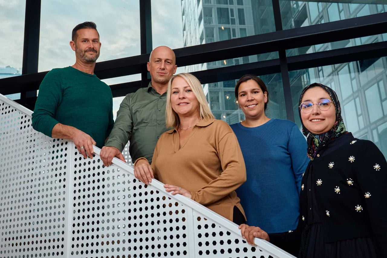 Five diverse professionals standing on a modern staircase in front of a glass-walled office building, posing for a group photo with friendly expressions.