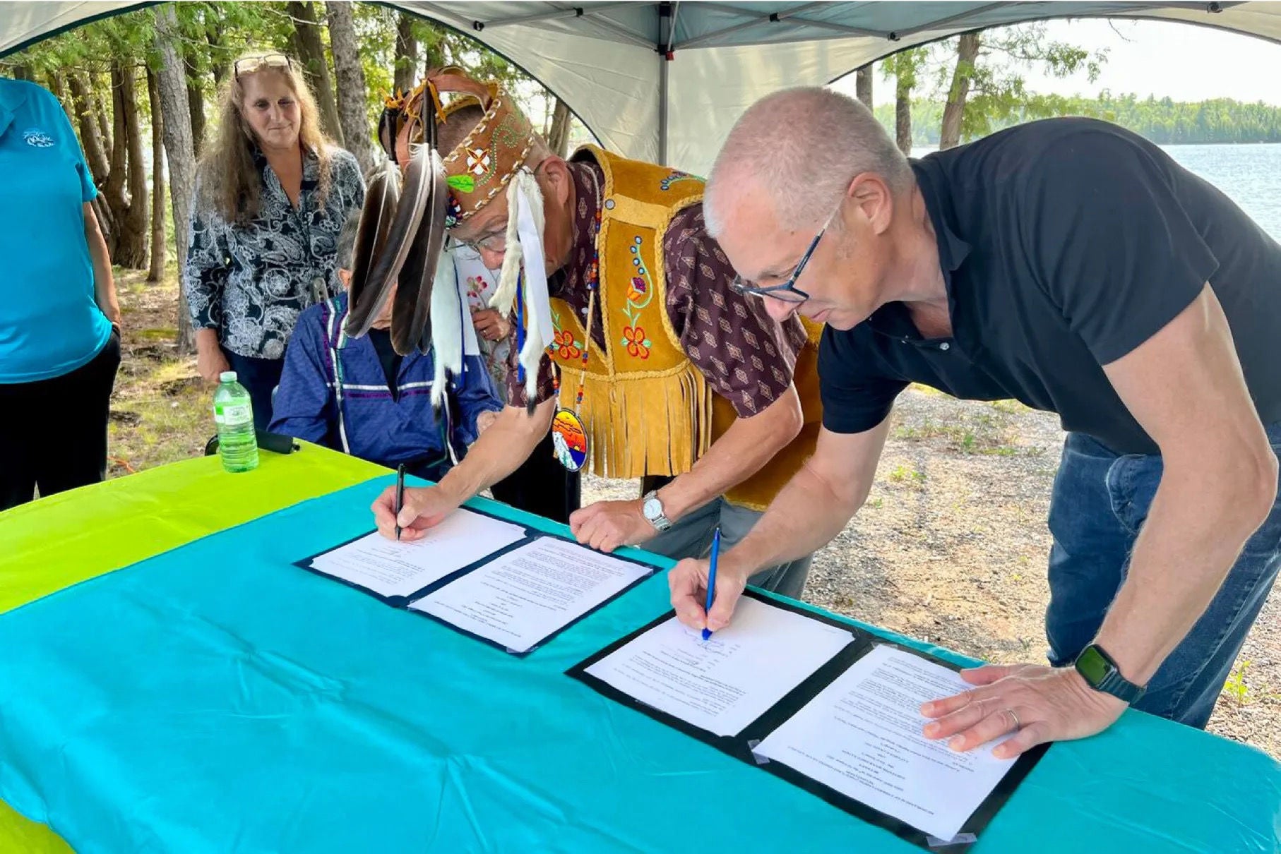 Two men signing documents; one is dressed in indigenous cultural attire and the other is dressed in casual business attire