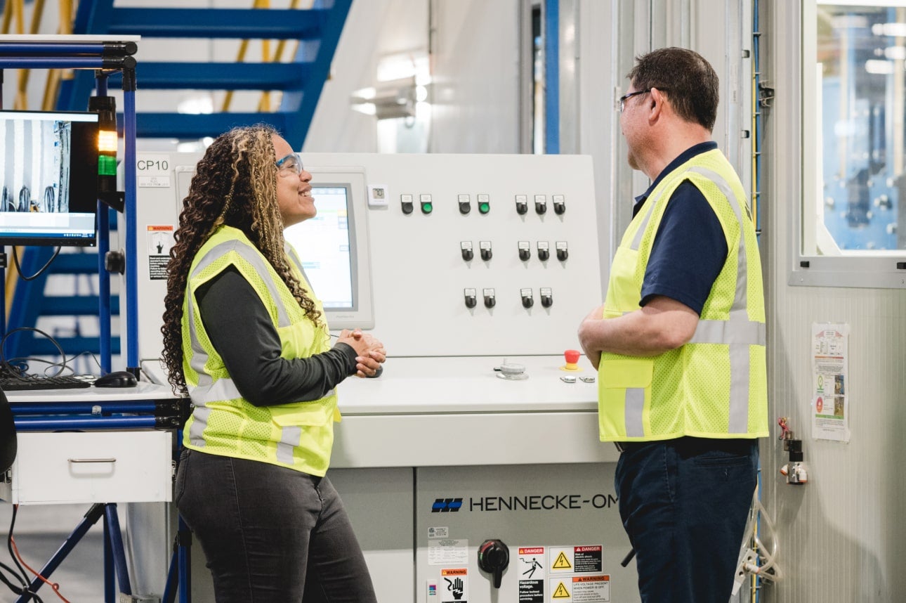 Two coworkers wearing safety vests conversing next to a digital machine in a modern industrial facility