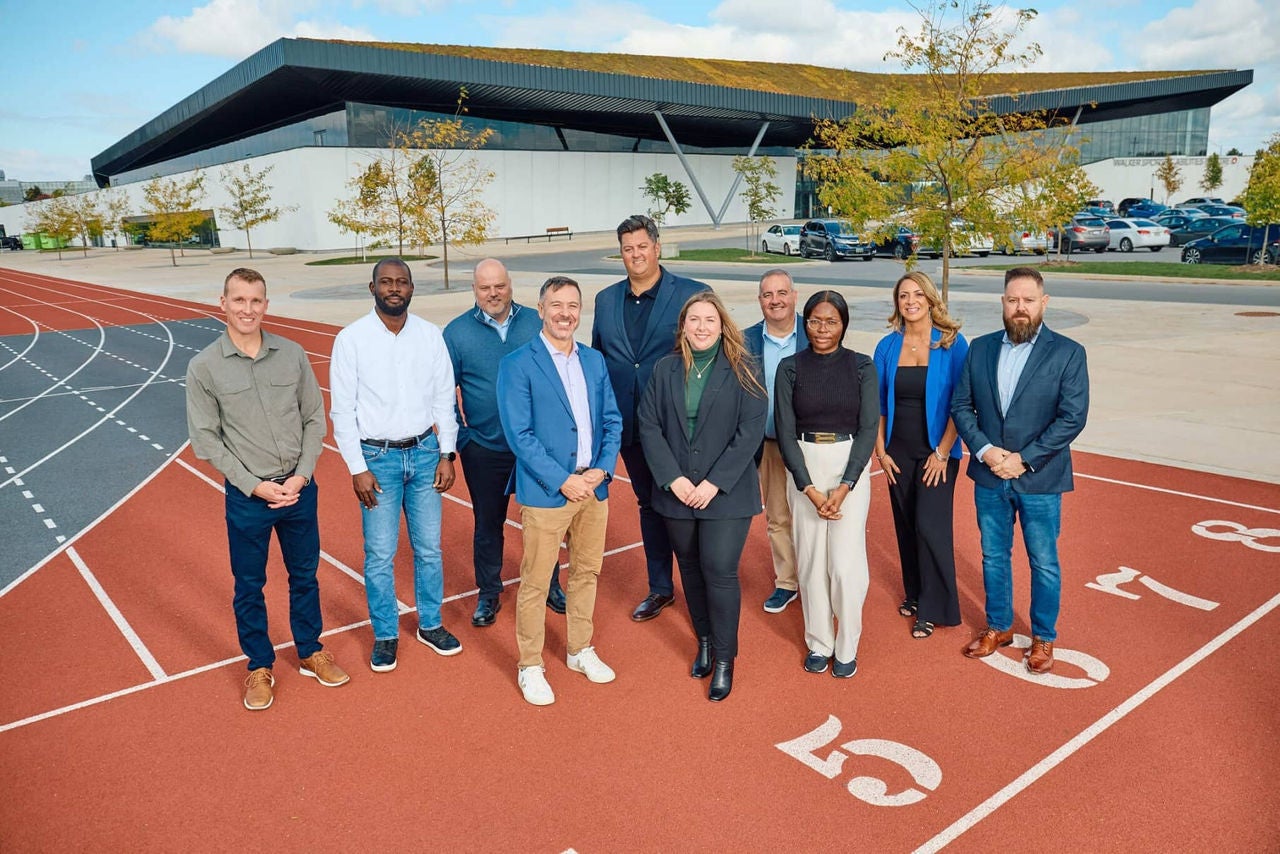 Ten professionals standing on a running track in front of a modern sports facility, posing confidently for a group photo on a sunny day.