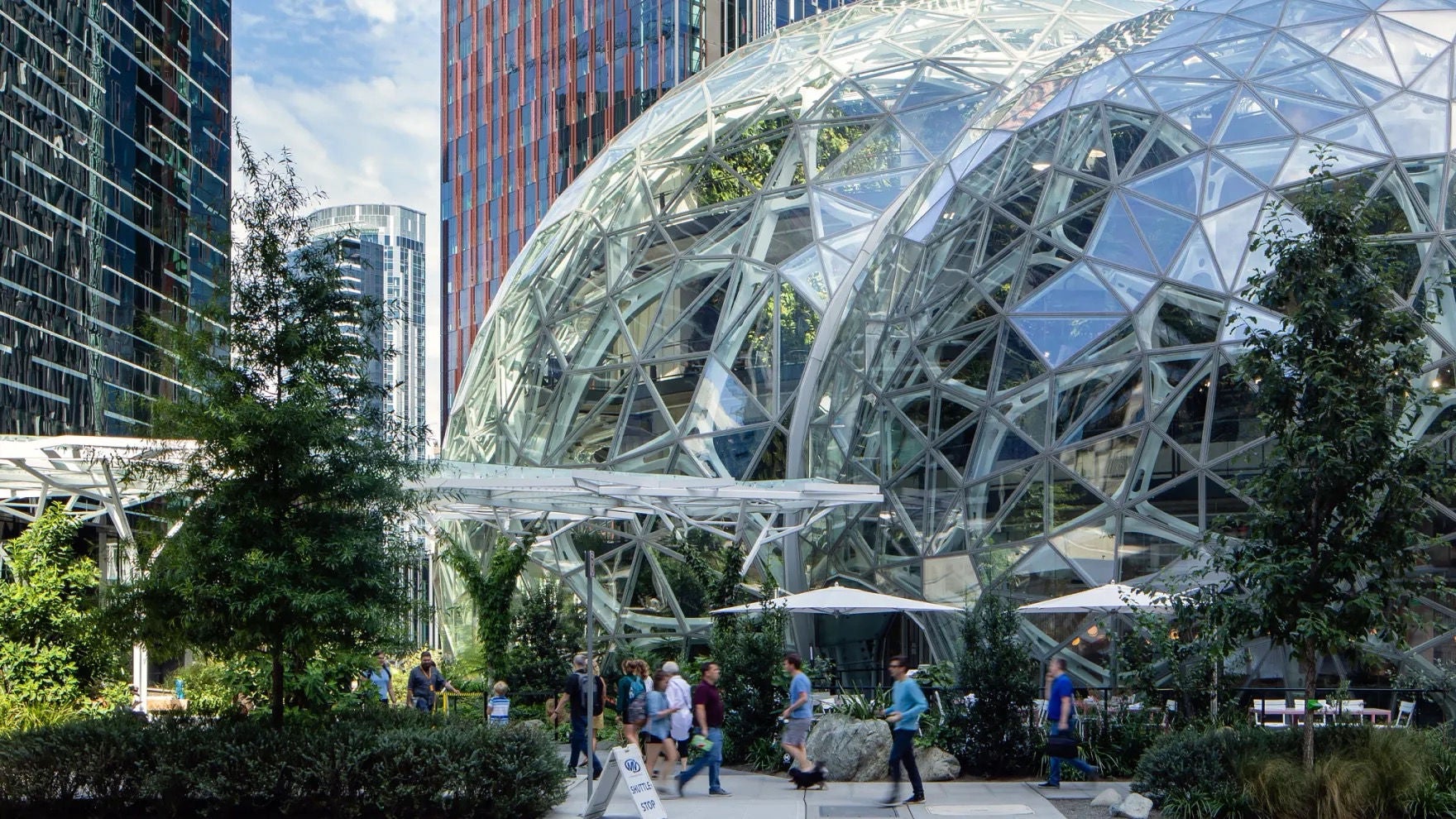 Exterior view of the Spheres from street level, with Seattle highrises in the background and people walking on the sidewalk in the foreground.